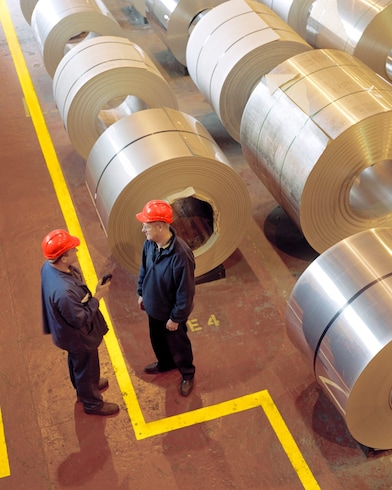 Two factory workers talking next to large rolls of steel