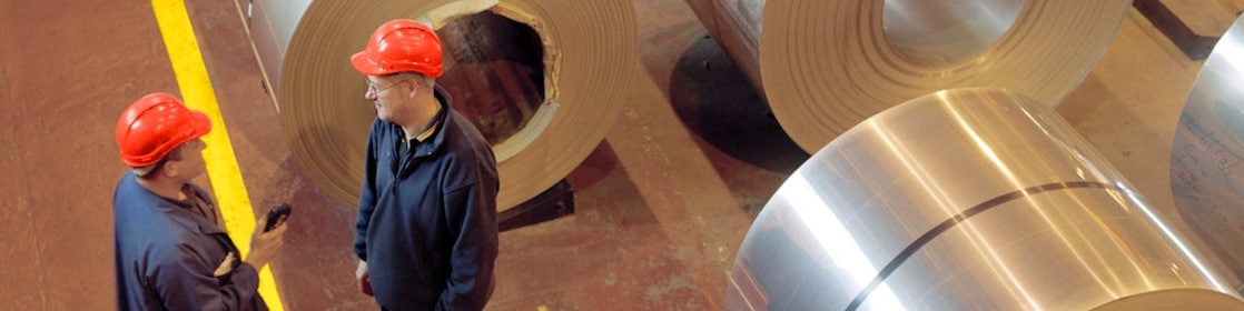 Two factory workers standing next to rolls of steel
