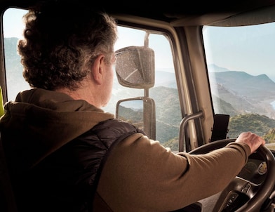 A truck driver steering along a coastal road with a scenic view of mountains and the sea.