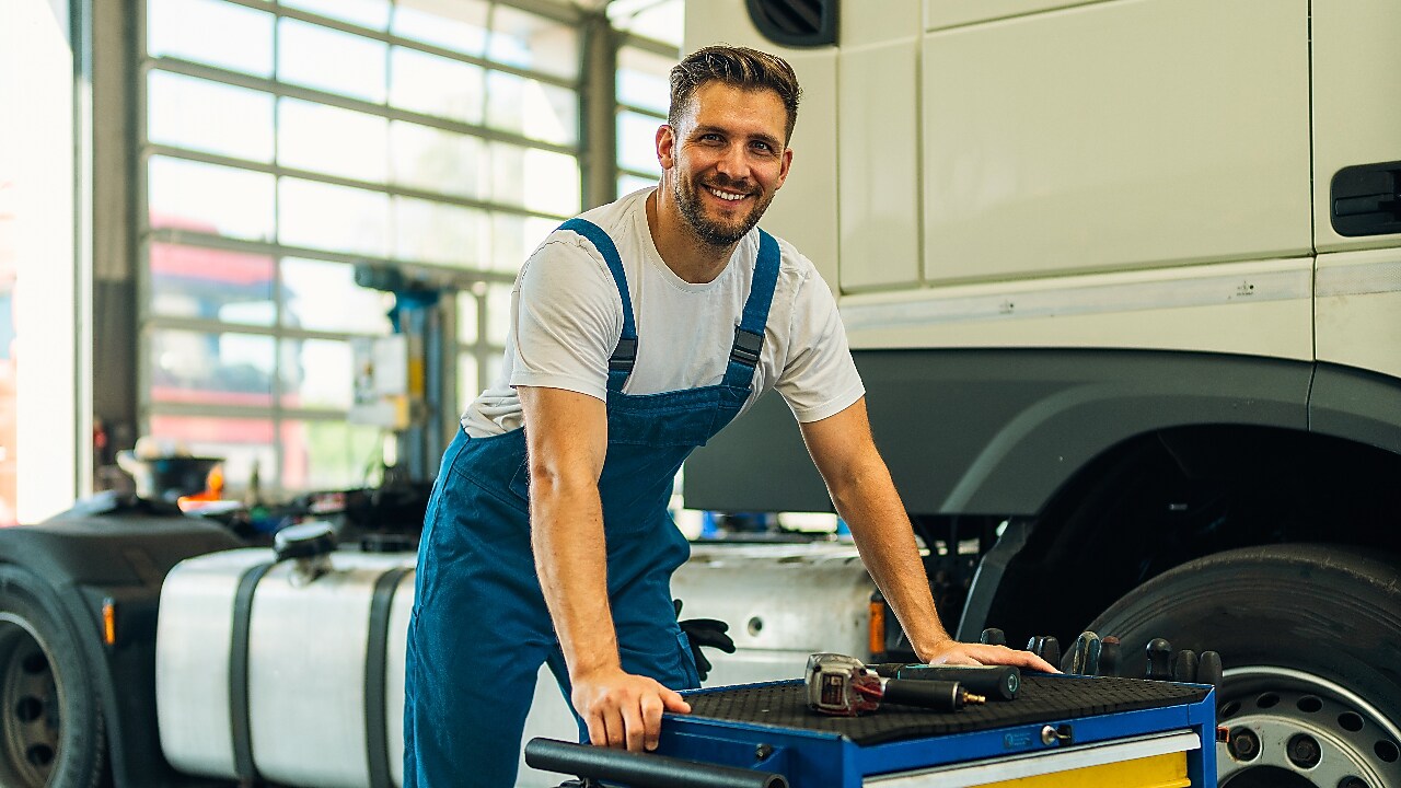 Ritratto di un tecnico positivo e sorridente con i suoi strumenti in piedi vicino al veicolo del camion in officina. Manutenzione e assistenza di veicoli industriali.