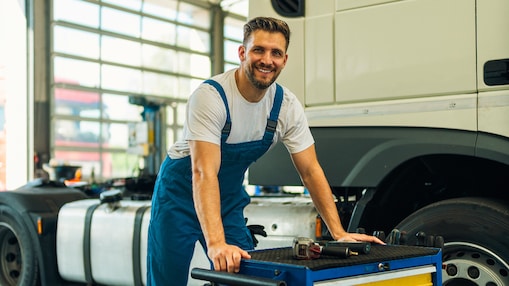 Ritratto di un tecnico positivo e sorridente con i suoi strumenti in piedi vicino al veicolo del camion in officina. Manutenzione e assistenza di veicoli industriali.